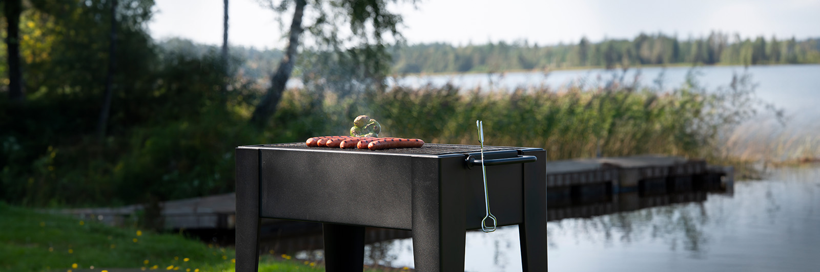 Outdoor park grill with sausages cooking, you can see a lake and trees in the background.